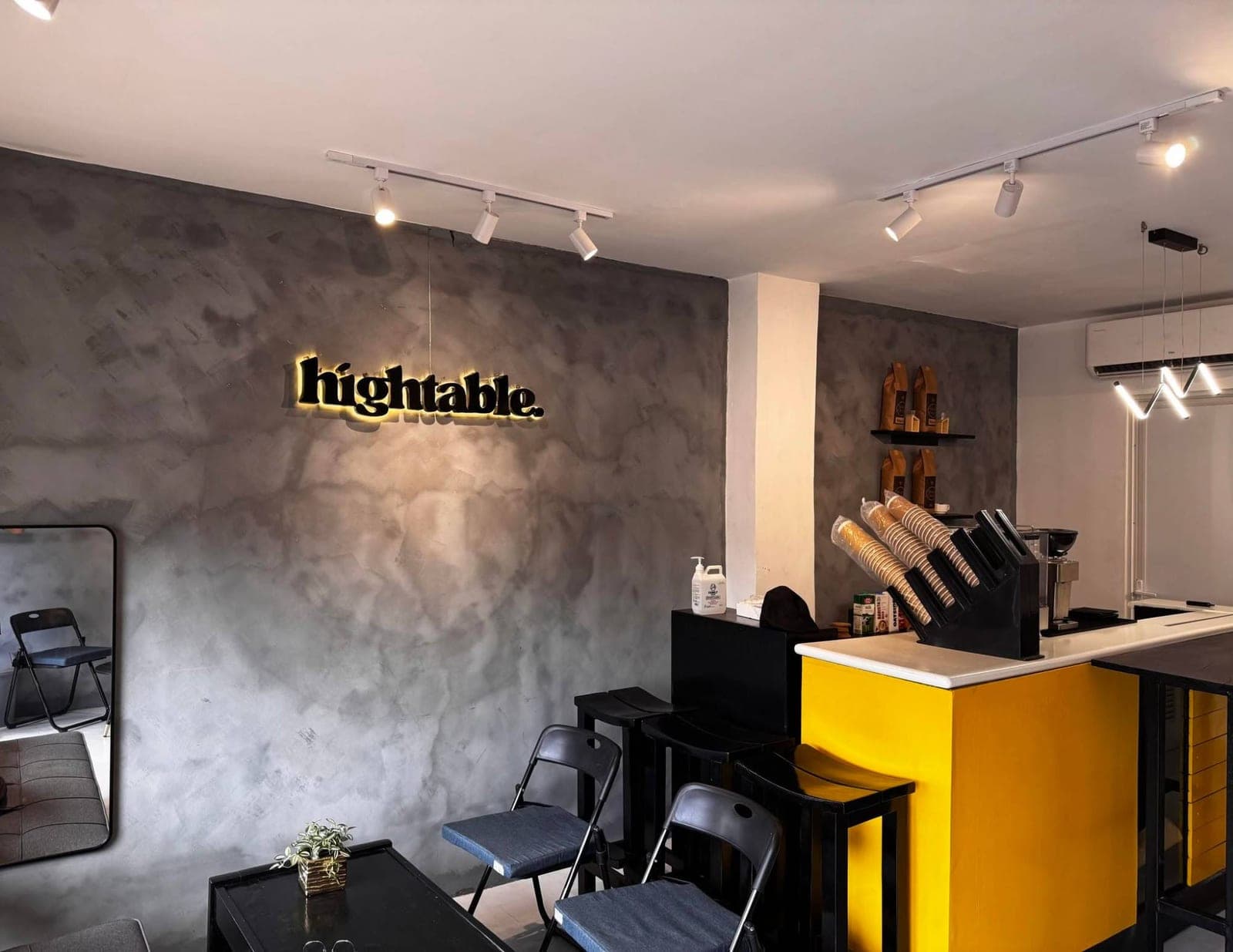Inside Hightable: a concrete wall with the backlit hightable sign, black stools and a yellow espresso bar